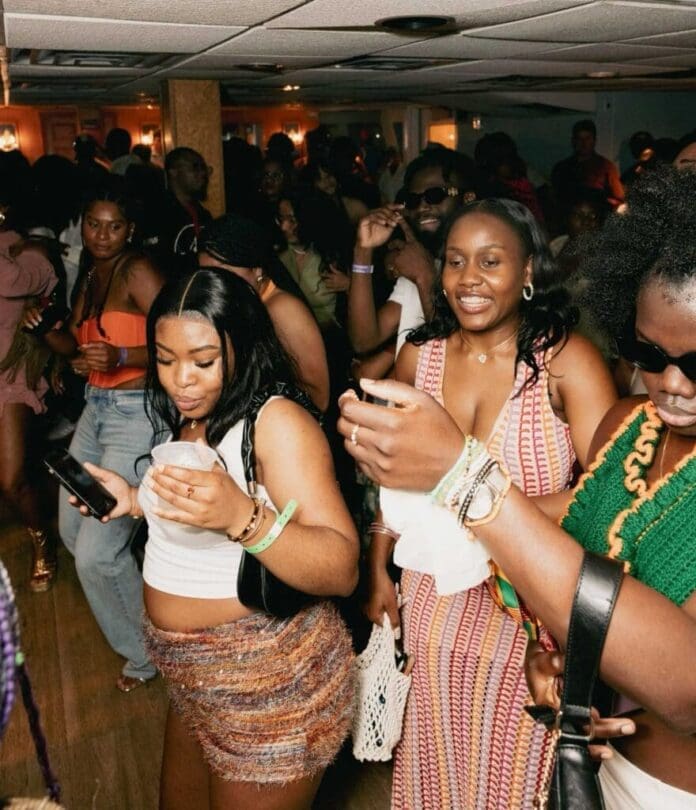 A group of people dancing and enjoying themselves at an indoor party, with several women smiling and holding drinks or phones, as Afrobeats music fills the air, celebrating African cultures.