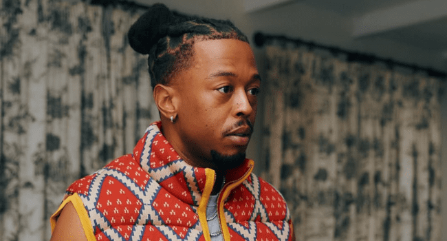 A man with braided hair and facial hair, reminiscent of DJ Zan-D’s era-defining run, is wearing a patterned red and yellow vest, standing indoors before patterned curtains.