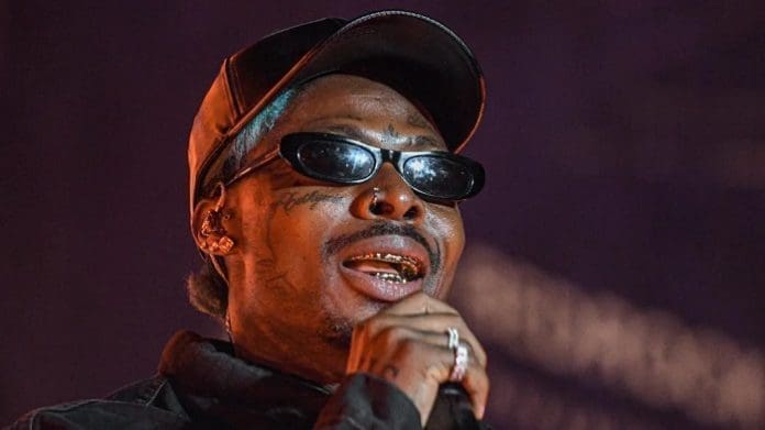 A person wearing a black cap, black sunglasses, and grills sings into a microphone against a dark background at the AfroFuture Festival in Accra.
