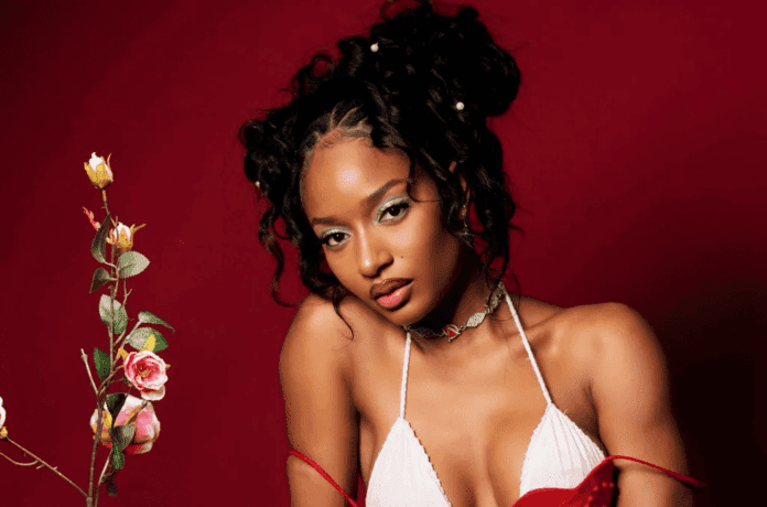 A woman with curly hair, wearing a white top and choker necklace, poses in front of a red background next to a stem with pink and yellow roses, capturing the vibrant energy often seen at the MOBO Awards.
