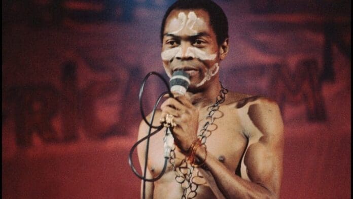 A shirtless African artist with body paint holds a microphone and stands on stage with a blurred red background, channeling the legendary Fela Kuti.