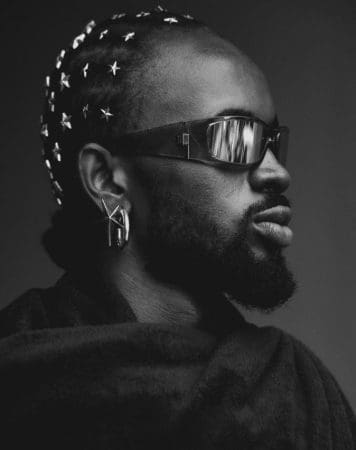 A person with braided hair adorned with star-shaped beads, wearing dark sunglasses and large earrings, is shown in a side profile against a dark background—radiating the cool energy of one of Apple Music's Biggest Artists.