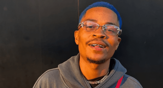 A man with blue hair, glasses, and a hoodie smiles while standing in front of a dark background, channeling Blue Pappi’s bold style from the new single “New Money.”.