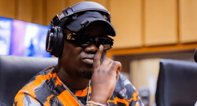 A person wearing headphones, a black cap, sunglasses, and an orange patterned shirt sits in a studio, channeling realness as they hold a finger to their lips—capturing the unique vibe of Blxckie's music success.