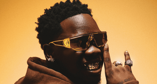 A man wearing dark sunglasses, a brown hoodie, diamond earrings, and rings, smiling and showing gold grills on his teeth, poses against a warm yellow background—a scene straight from a Romantic Teaser for Blxckie's 4LUV 2.