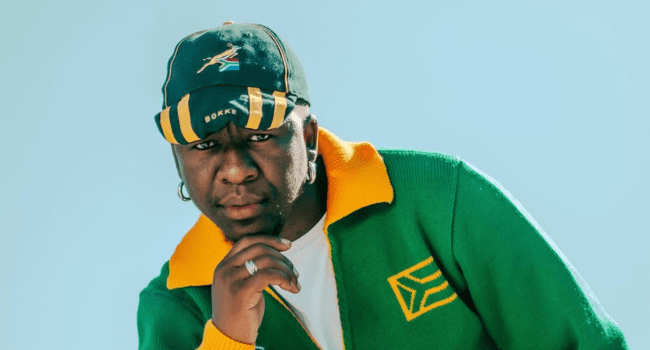 A person in a green and yellow jacket and matching cap with South African flag colors looks at the camera, resting their chin on their hand against a clear sky—capturing a thoughtful farewell vibe reminiscent of Priddy Ugly.
