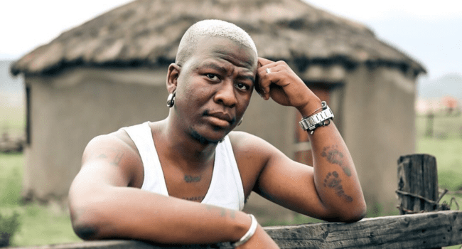 A person with short blond hair and tattoos, reminiscent of rapper Bravo Le Roux, leans on a wooden fence in a white tank top, with a round thatched-roof hut in the background.
