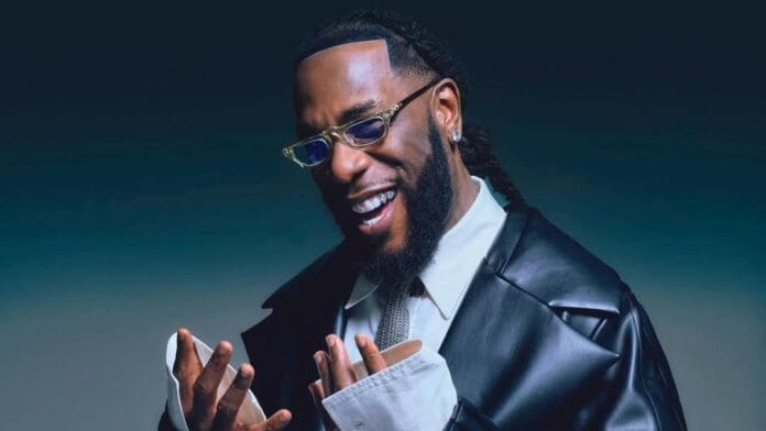 A man wearing glasses, a black leather jacket, and a tie smiles while looking at his hands against a dark background, reminiscent of Burna Boy celebrating his first song in The Guardian Nigeria.