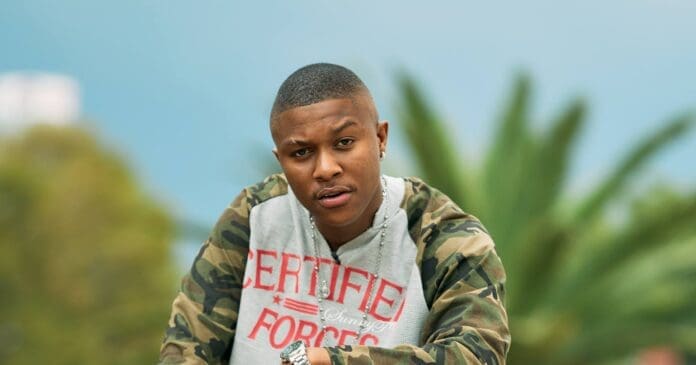 A person wearing a camouflage jacket and a gray "CERTIFIED" hoodie looks at the camera, embodying Pitori Culture, with trees and a blurred background behind them.