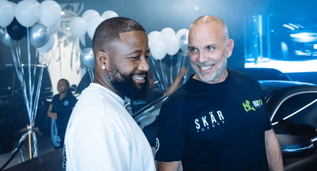 Two men stand smiling in a showroom decorated with balloons and cars, with one wearing a white shirt and the other in a black "SKÄR" shirt, posing next to the Cassper Maybach GLS 600 after its Stealth-Luxury Upgrade.