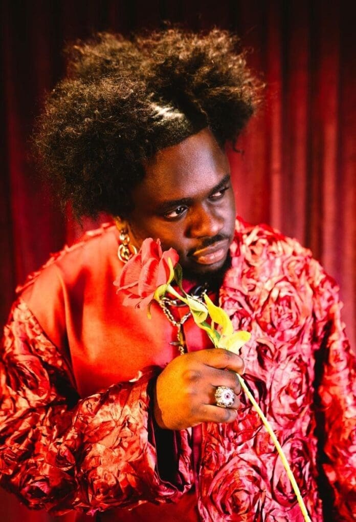 A person in a textured red outfit holds a red rose close to their face, standing before a burgundy curtain—capturing the moody vibe of Coblaze’s Piece Of Me EP featured by The Culture Custodian.