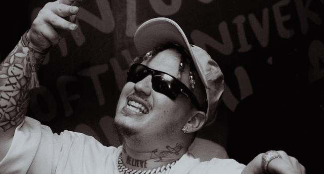 A trailblazer in style, Costa Titch smiles and gestures with his hand, wearing sunglasses, a cap, and jewelry in front of a wall with large text. The black-and-white image captures his vibrant spirit.