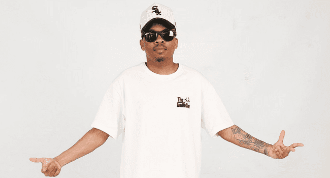 Man wearing a white t-shirt, white baseball cap, and sunglasses stands with arms slightly outstretched against a plain background, channeling the cool vibes of Dj Clen’s “Rollin’.”.