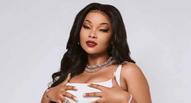 Woman with long black hair and red lipstick, wearing a white top and silver jewelry, poses with hands on her chest against a plain light background—capturing DJ Fae Fae's strength after her hardest year and life change.