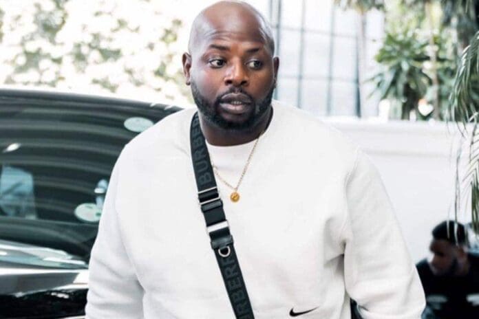 A man in a white sweatshirt with a black Burberry strap stands outdoors near a car, with greenery and a glass building behind him—reminiscent of DJ Maphorisa amidst ongoing exploitation allegations.