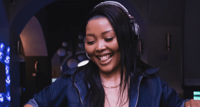 DJ Miss Pru, wearing headphones and a denim jacket, smiles while standing indoors—possibly DJing her hit track "Ameni" or celebrating a radio milestone.