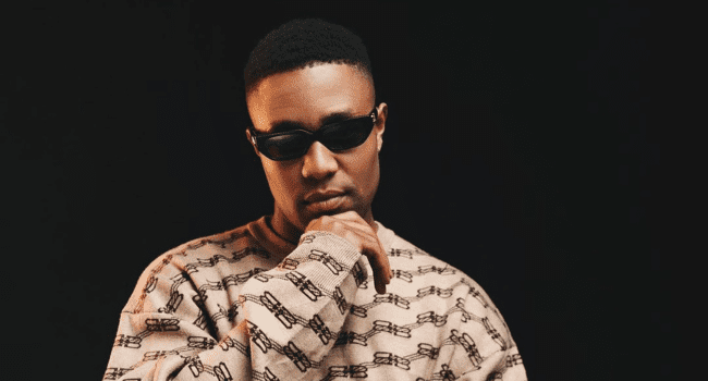 A person wearing dark sunglasses and a patterned sweater stands against a black background, resting their chin on their hand—waiting for ticket requests to the DJ Speedsta Gunna show.