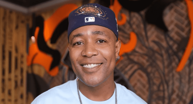 A man wearing a blue baseball cap and light blue shirt smiles at the camera with abstract wall art in the background, capturing the vibrant energy of a DJ Speedsta festival set at the Milk + Cookies Festival.