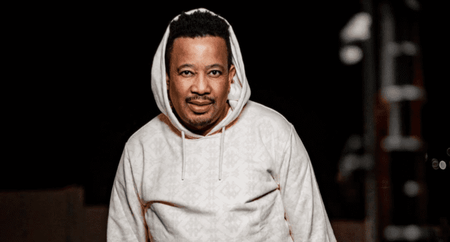 A man wearing a light-colored hoodie stands outdoors at night, facing the camera with a neutral expression—channeling DJ Zan-D's Hip Hop vibe.