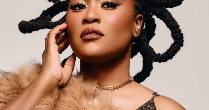 A woman with coiled locs poses with her hand on her face, channeling an Afrobeats vibe in a fur stole, gold necklace, and black top against a neutral background.