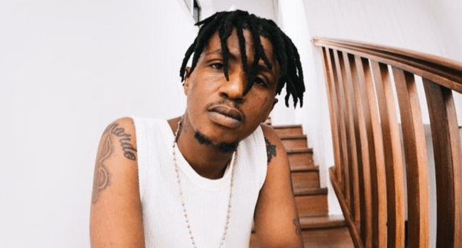 A man with short dreadlocks and tattoos on his arms wears a white sleeveless shirt and necklace, sitting on wooden stairs next to a railing—reminiscent of scenes from an Emtee music video like WAKE UP / WAR.