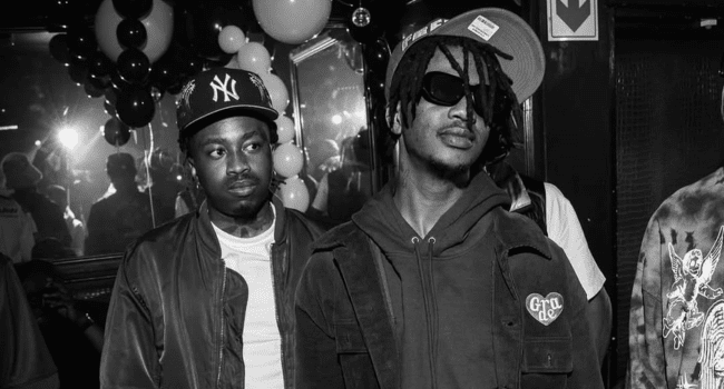 Two men stand indoors at a Trap House Party; one wears a hoodie and sunglasses, the other a NY cap and jacket. Balloons and people fill the background at this secret location.