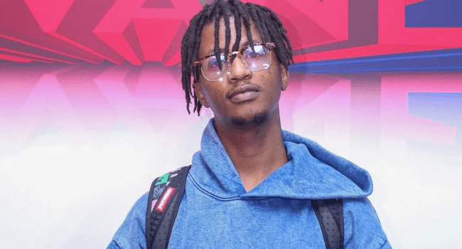 A person wearing glasses, a blue hooded sweatshirt, and a black backpack stands in front of a colorful geometric background, their expression hinting at pain as if they've been taken advantage of.