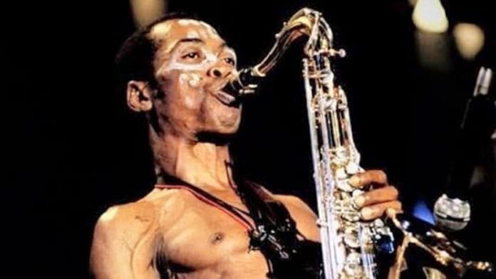 A shirtless man with face paint showcases his musicianship as he plays a saxophone on stage under bright lights, echoing the energy of Fela.