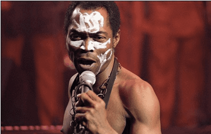 A man with white face paint sings into a microphone on stage, channeling Afrobeats vibes as he wears a necklace and dark clothing against a reddish background.
