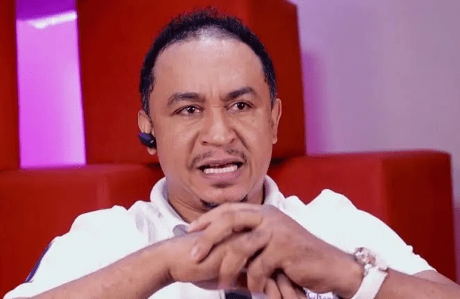 A man wearing a white shirt and earpiece, resembling Daddy Freeze, sits with his hands clasped, speaking against a background of red and purple tones.
