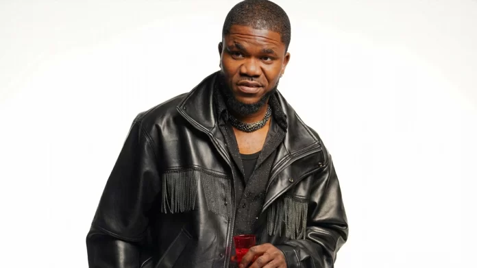 A man wearing a black leather jacket and a black shirt holds a red drink, standing against a plain white background—a style often seen among leading diaspora artistes in the Afrobeats industry.
