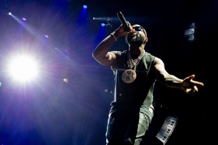 A rapper wearing sunglasses and a large pendant performs Afrobeats on stage under bright lights, holding a microphone and reaching toward the audience, capturing the energetic vibe of West Africa’s Detty December.