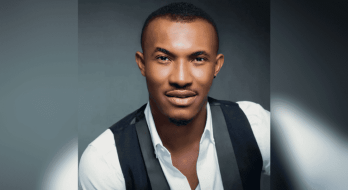 A man with short hair and a trimmed beard wears a white shirt and black vest, looking directly at the camera against a dark background—reminiscent of Nollywood actor Gideon Okeke or rising Afrobeats stars.