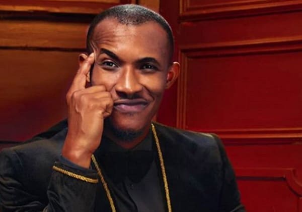A man in a black suit with gold trim touches his temple and smiles, seated in front of a wooden paneled background, channeling the confident style seen in Nollywood films.