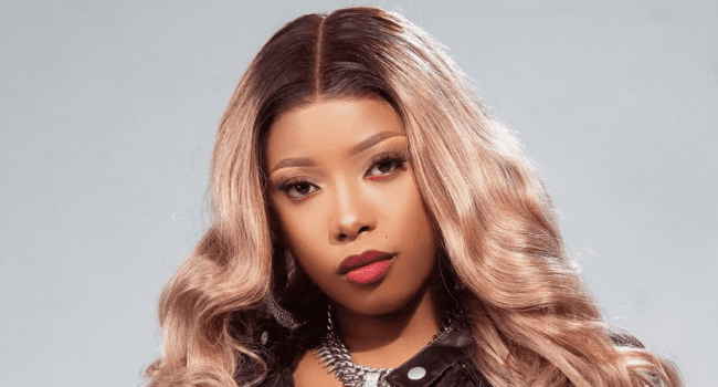 A woman with long, wavy, light brown hair and red lipstick looks at the camera against a plain gray background. Channeling Gigi Lamayne’s style, she wears a black outfit and a silver chain necklace.