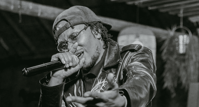A person wearing glasses, a cap, and a leather jacket holds a microphone and performs, extending one hand forward—perhaps searching for missing songs in the Ginger Trill catalogue.