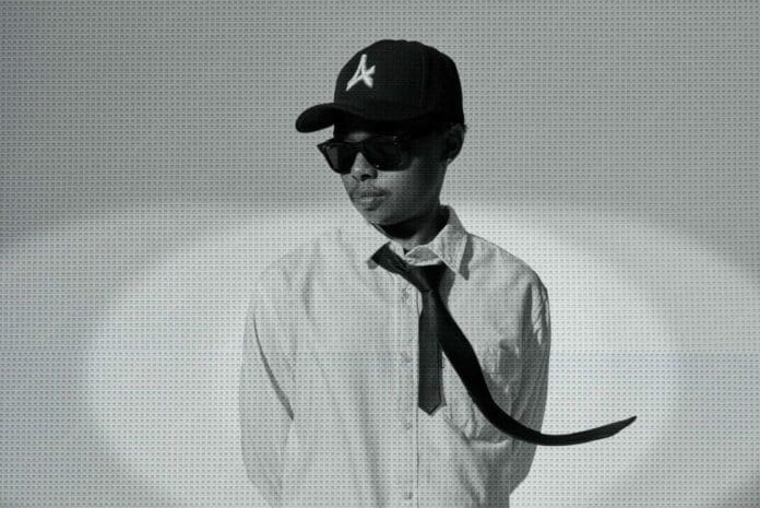 Person wearing a white shirt, black tie, black sunglasses, and a black cap stands against a plain backdrop; the loose tie is blown sideways, channeling the cool vibes of A-Reece's "Before I Overthink" in this midnight drop look.