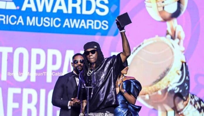 A musician in sunglasses and a black cap raises an AFRIMA award on stage at the Africa Music Awards, with two people standing behind him, celebrating the rise of Afrobeats.