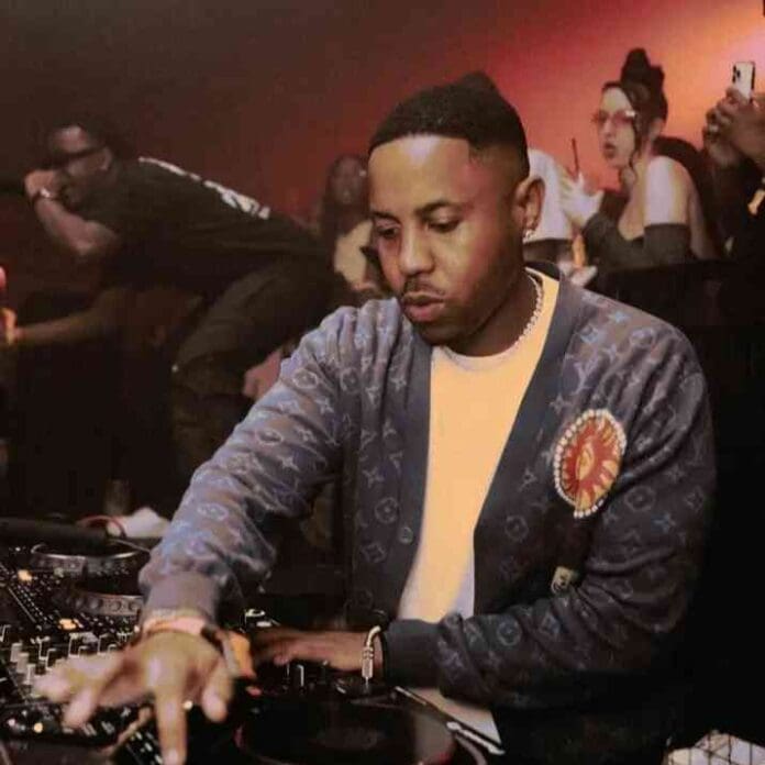 A man in a patterned jacket operates DJ equipment in a dimly lit club, spinning an energetic Amapiano Mix as people dance and cheer in the background.