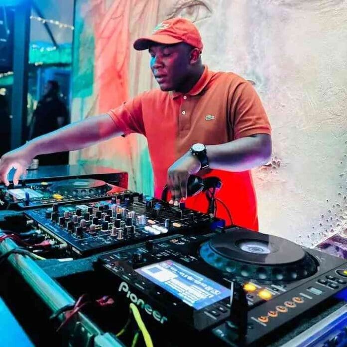 A DJ wearing a red cap and brown polo shirt operates a professional DJ mixer and turntables in a colorful indoor setting, spinning tracks like "Nilavha Wena" by Busta 929 and Dj Jaivane.