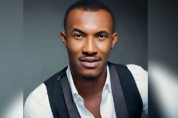 A man with short hair and slight facial hair, wearing a white shirt and dark vest, looks into the camera with a neutral expression against a dark background, reminiscent of Gideon Okeke from the film industry.
