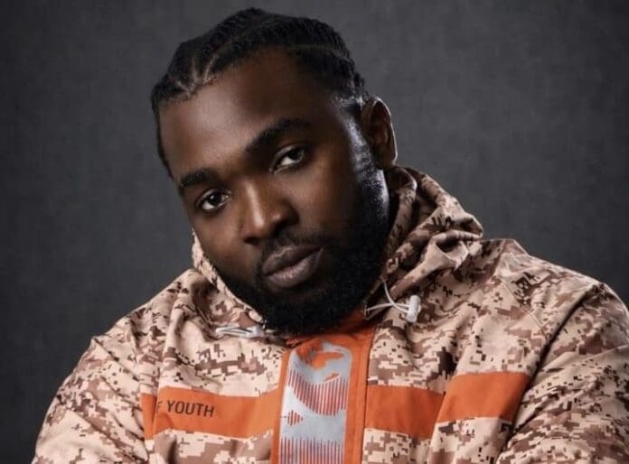 A man with braided hair and a beard wears a patterned beige jacket labeled "YOUTH" and looks directly at the camera against a dark background, channeling Afrobeat style.