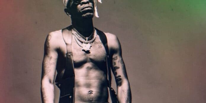 A shirtless man wearing a bandana, necklaces, and suspenders stands facing forward with tattoos visible on his arms and torso, channeling the bold style of Shatta Wale and Echoes of The African Ghetto.