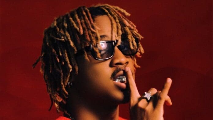 Person with blond dreadlocks and sunglasses holds a finger to their lips in a shushing gesture, wearing a red shirt and jewelry against a red background, channeling Afrobeats vibes for CupidSZN.