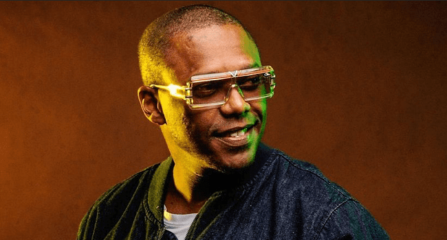 A man wearing large, square glasses and a dark jacket looks to the side and smiles against a brown background with green lighting, giving off DJ Switch vibes.