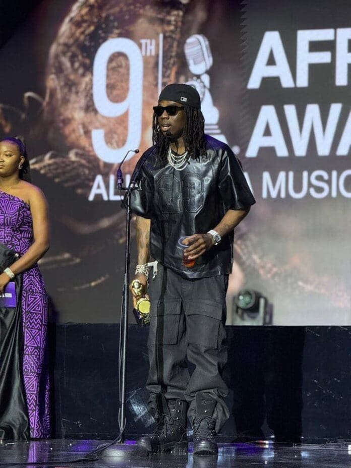 A man in black leather clothes and sunglasses stands on stage holding a microphone and an award, with a woman beside him at the 9th African Music Awards, celebrating the vibrant scene of African music.