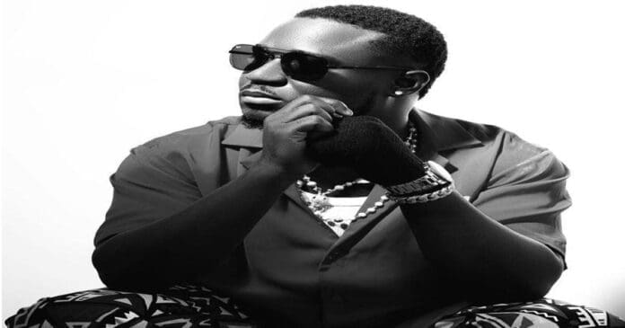 A man wearing sunglasses, a short-sleeved shirt, patterned pants, jewelry, and a wristwatch poses with his hand on his chin against a white background, capturing the fresh vibe of Ichaba’s new music for 2026.