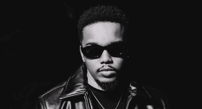 A man wearing dark sunglasses and a leather jacket looks at the camera against a plain black background, channeling the intense energy of The People Are Burning by Jay Jody, ready for his next LIVE Concert.