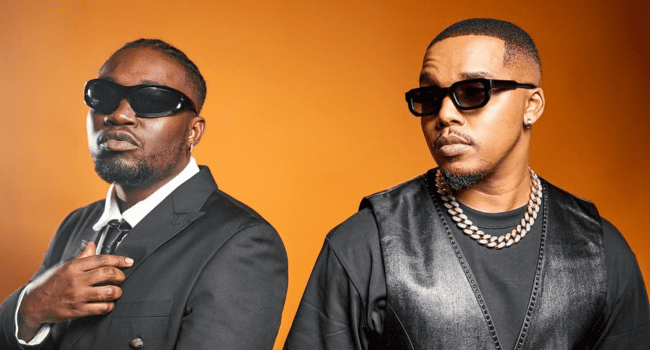 Two men, channeling Manu WorldStar vibes, pose in dark sunglasses and black outfits against an orange background. One wears a suit and tie; the other rocks a chain necklace and sleeveless vest, embodying The People Are Burning energy.