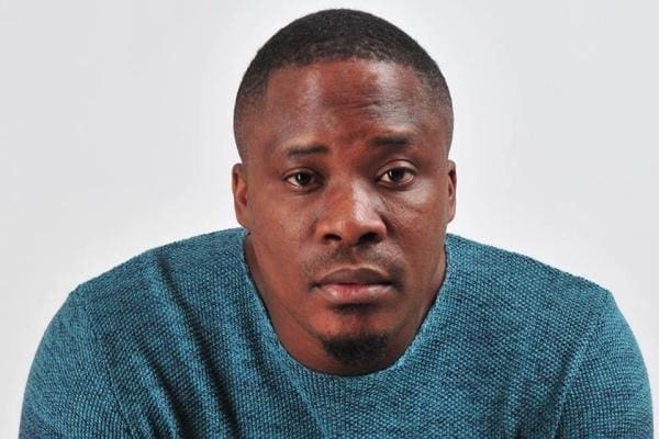A man in a teal sweater sits against a plain white background, looking directly at the camera with a neutral expression, reminiscent of Jaywon's reflective style noted in The Nation Newspaper’s coverage of his music impact.